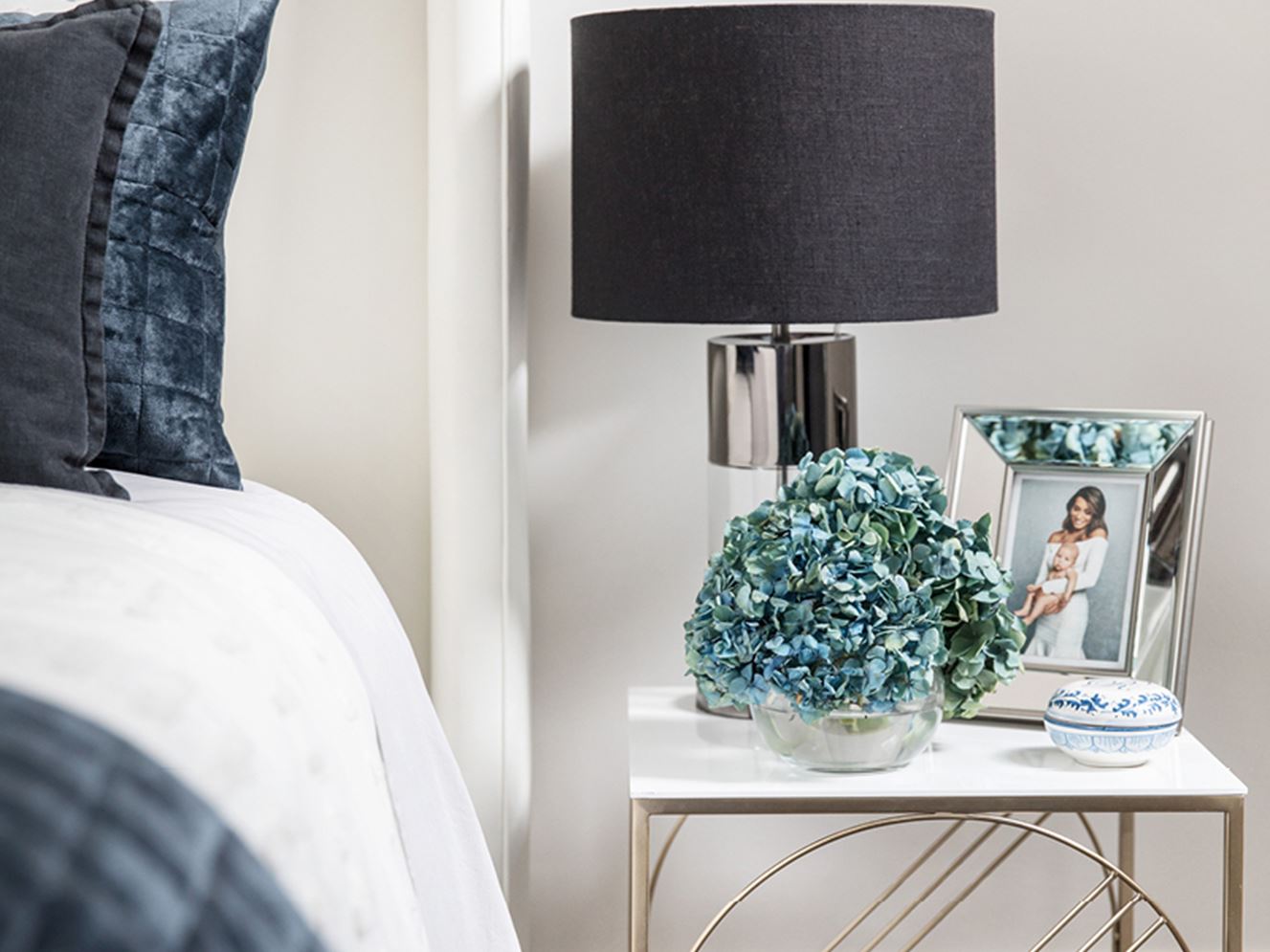Brushed brass bedside table with marble top, with blue hued flower arrangement and mirror edged photo frame.